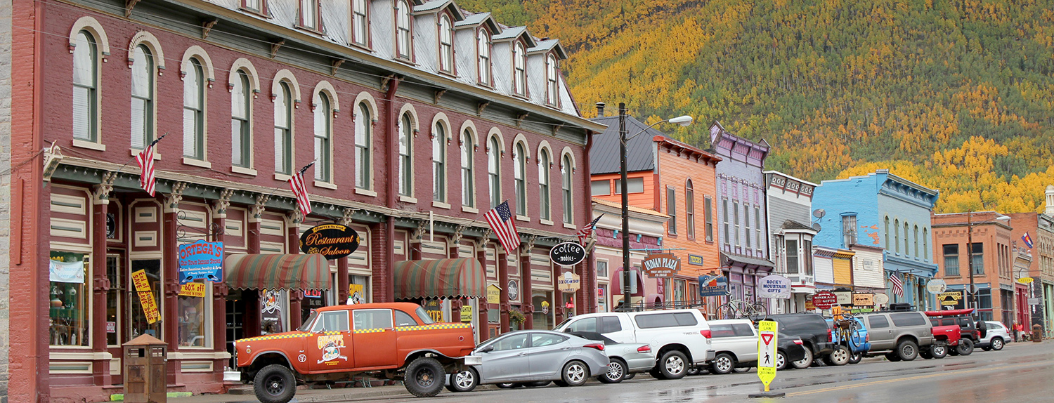 Main street in Silverton, with cars parked in front of a line of Victorian-style brick or wood buildings and a hillside of golden aspen trees in the background