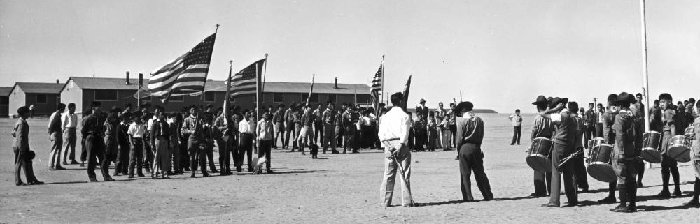 A group of Japanese American Boy Scouts stand in formation on the grounds of Amache on May 30, 1943. The barracks where they were forcibly detained are in the background. 