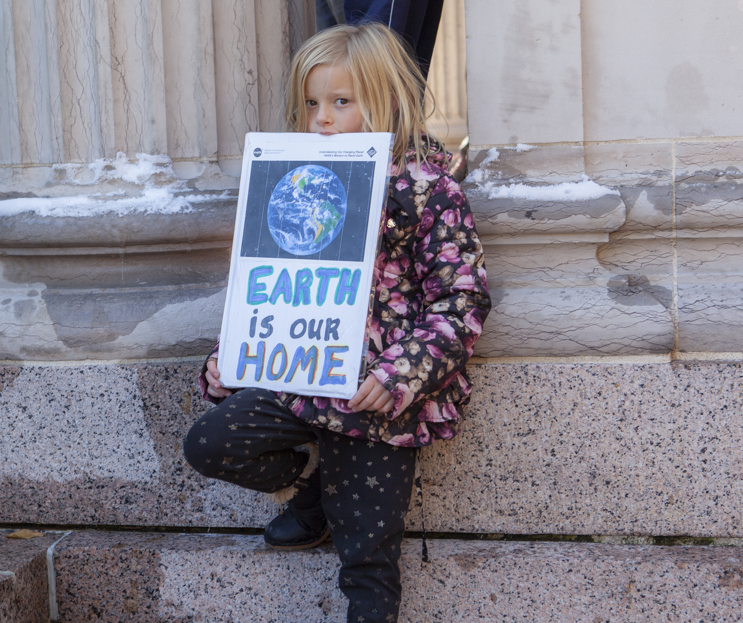 Girl on the steps of the capitol holds a sign that says Earth is Our Home