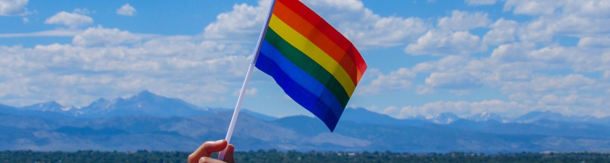 A rainbow pride flag waving in front of a mountain backdrop on a partly cloudy day