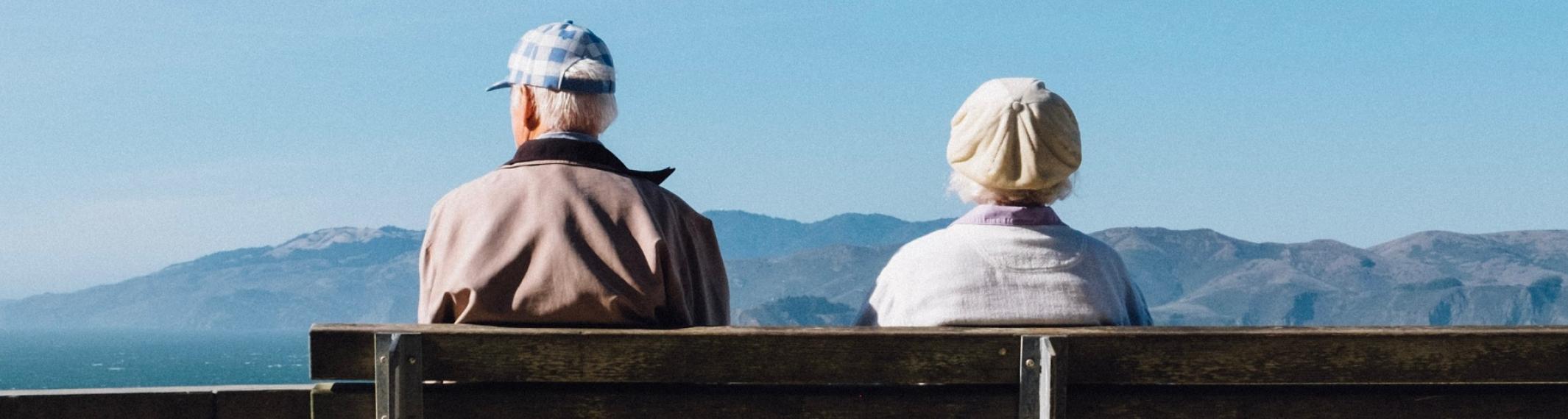 An older couple sits on a bench overlooking a mountain view.