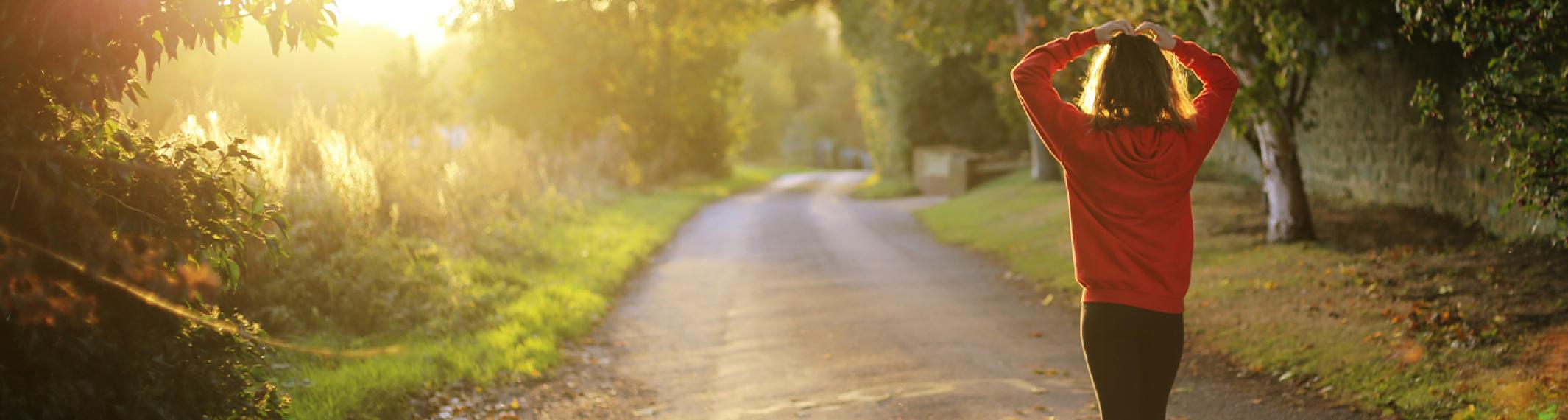 A woman in a red jacket standing at the edge of a dirt road. Her back is to the camera.