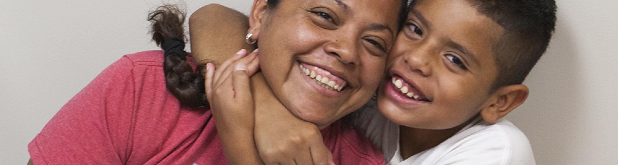 A mom in a red T-shirt smiling and leaning to the right toward her eight-year-old son as he smiles and wraps his arms around her neck