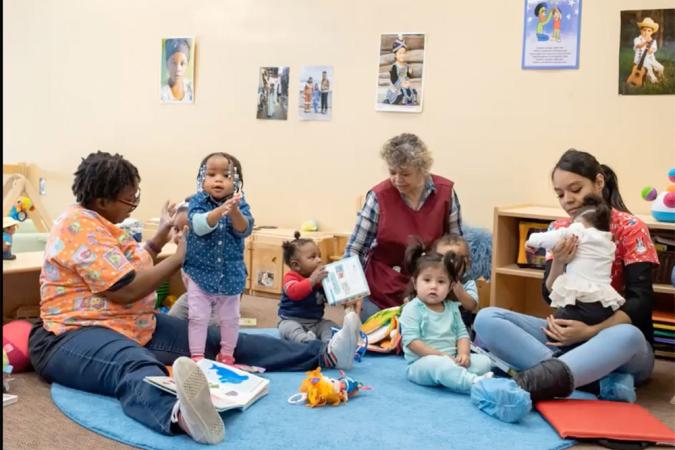Three women and three toddlers sitting on the floor in a child care room.