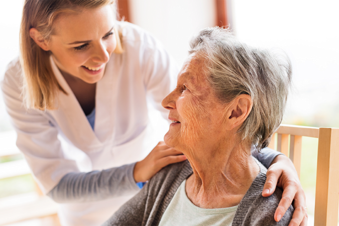 A young woman in a white medical coat smiling at an older woman while she places her hands on the older woman's shoulders