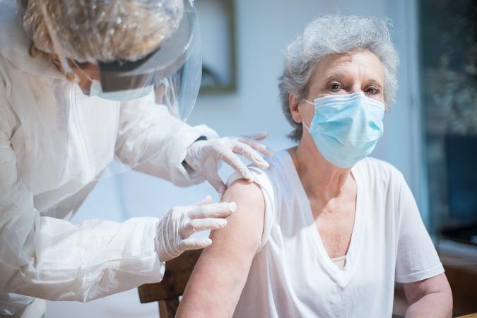 A nurse in a face shield and protective coat touches the arm of a woman with gray hair who is wearing a face mask, as she prepares to give a vaccine.
