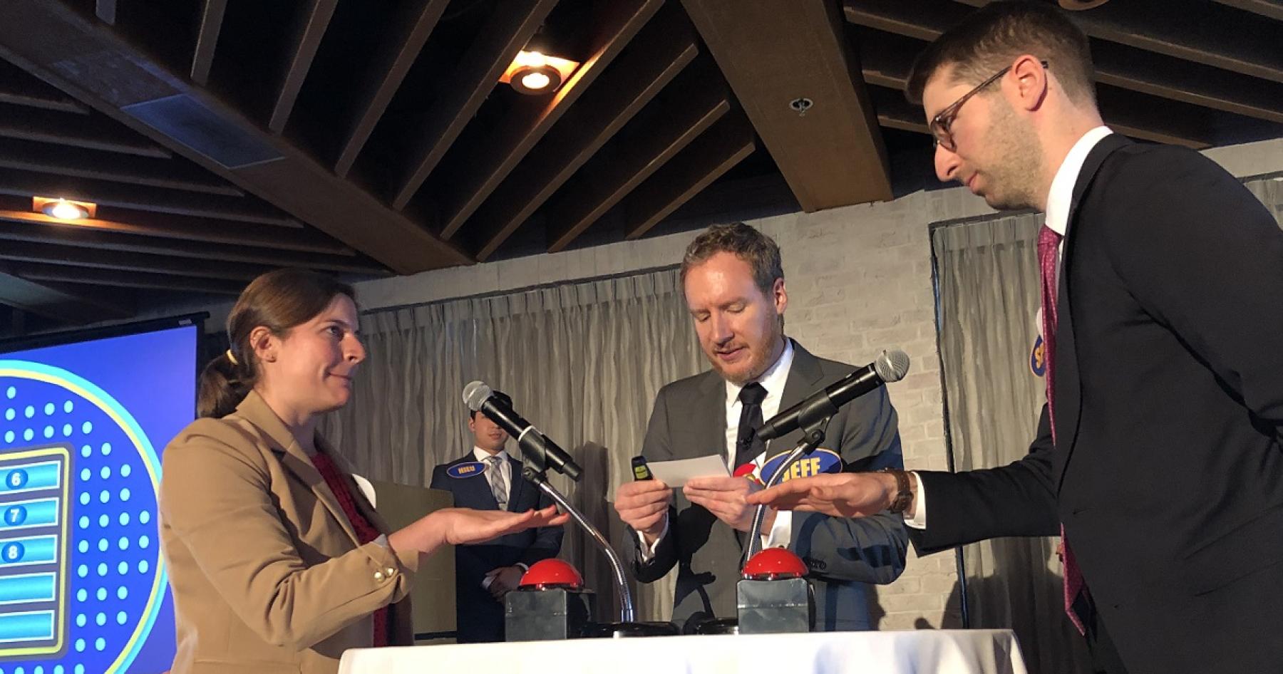 Nina Bastian and Spencer Bud hold their hands over game show buzzers, as Jeff Bontrager reads a question.