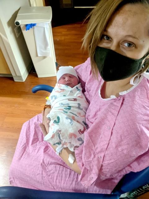 A woman in a pink hospital gown and face mask sitting down holding a newborn