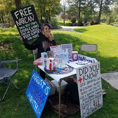 A woman sits at a table in a park holding a sign reading "Free Narcan" and distributing informational flyers.