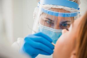 A dentist in a paper mask and clear face shield uses a dental pic on an adult woman, whose face is blurred in the foreground